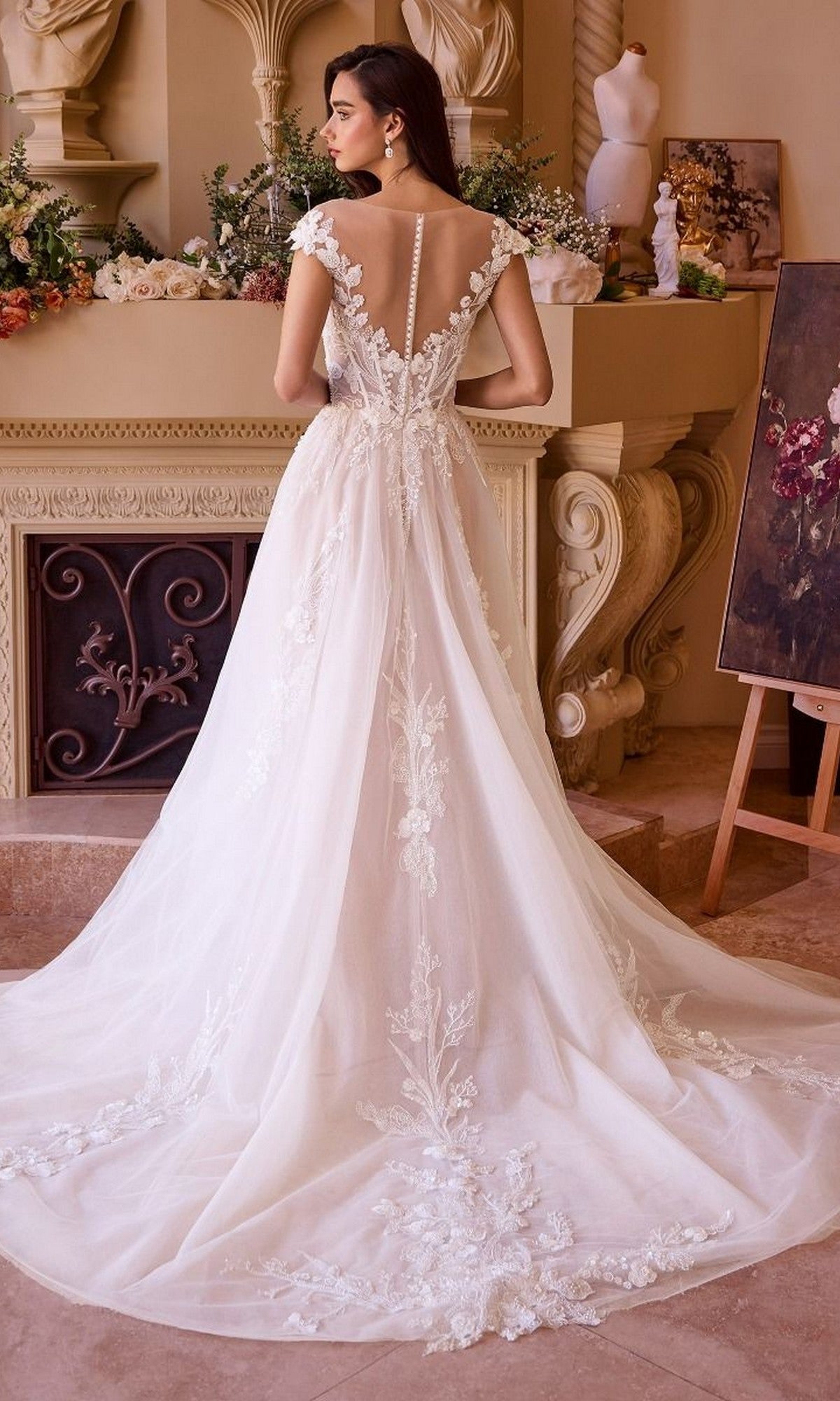 A woman in the Andrea and Leo WL038 long wedding dress with cap sleeves, lace applique, and a sweeping train stands indoors facing a decorated fireplace.
